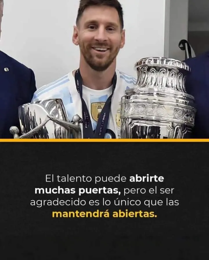 Se ve a Lionel Messi sonriendo con una copa en la mano. La copa es de color dorado y tiene asas. Messi lleva una camiseta azul y blanca a rayas. Tiene el pelo corto y negro. Está en un lugar que parece ser un estadio de fútbol. Hay mucha gente en las gradas. Todos están contentos y celebran la victoria de Messi.